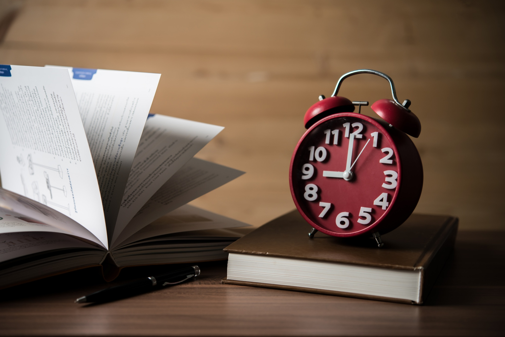 A classic red alarm clock sitting on a stack of books beside an open book and a pen