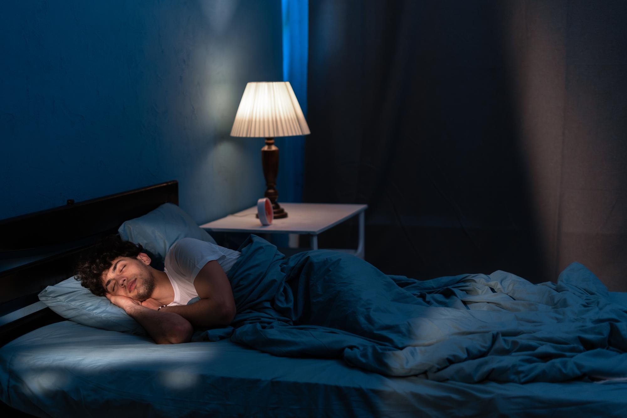 A cozy dim bedroom at night with someone asleep on their side, soft linen bedding and warm lamp glow